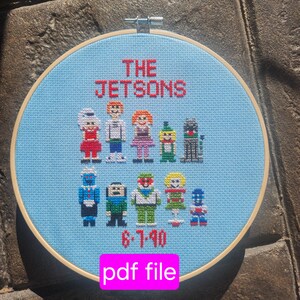May include: A cross-stitch hoop art piece featuring the Jetsons family. The design includes the words "THE JETSONS" in red, along with pixelated characters and the date "6-7-90". The background is light blue, and the hoop is made of wood.