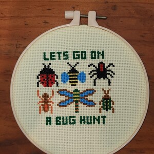 May include: A cross-stitch embroidery hoop featuring a cream-colored fabric with a design of various bugs and the text "LET'S GO ON A BUG HUNT" in green. The bugs include a ladybug, bee, spider, dragonfly, and beetle.