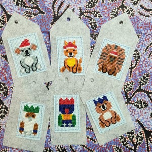 May include: A collection of five gray felt gift tags, each featuring a cross-stitched animal design. The animals include a koala, a kangaroo, a lion, a koala, and a colorful creature, all wearing festive hats. The tags have a small hole at the top.