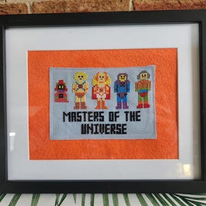May include: Framed cross-stitch art featuring characters from Masters of the Universe. The artwork is set against an orange background within a black frame. The characters are depicted in vibrant colors, with the text "MASTERS OF THE UNIVERSE" below.