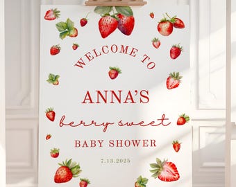 May include: A white sign on a wooden easel with red text and strawberry illustrations. The sign reads "Welcome to Anna's berry sweet Baby Shower 7.13.2025". The strawberries are red with green leaves.