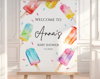 May include: A wooden easel displays a baby shower sign. The sign features watercolour illustrations of colourful ice lollies and the text "Welcome to Anna's Baby Shower 7.12.2025". The sign is decorated with confetti.