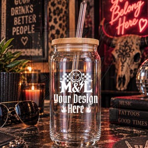 May include: A clear glass tumbler with a bamboo lid and a glass straw. The front of the glass features a white graphic print with a smiley face, a lightning bolt, and the text M&L Your Design Here, accented by checkerboard and flame icons.