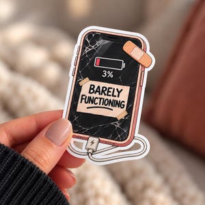 May include: A die-cut sticker of a smartphone with a cracked screen, a low battery icon at 3%, and a beige adhesive bandage on the corner. A piece of tape on the screen displays the text BARELY FUNCTIONING. A charging cable is plugged into the base.