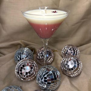 Könnte beinhalten: Eine zweifarbige rosa und weiße Kerze in einem Martini-Glas mit einem einzelnen roten Blütenblatt darauf. Die Kerze ist von silbernen und rosafarbenen Discokugel-Ornamenten umgeben.