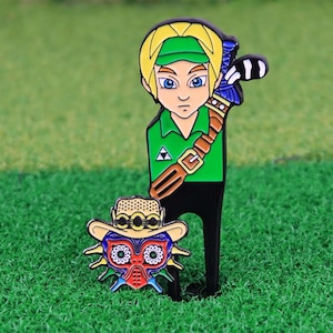 Peut inclure: Un outil de réparation de divot de club de golf en métal vert et noir représentant un personnage de dessin animé aux cheveux blonds, une tunique verte et une ceinture marron. Un deuxième marqueur de balle de golf en forme de masque coloré plus petit est également inclus.