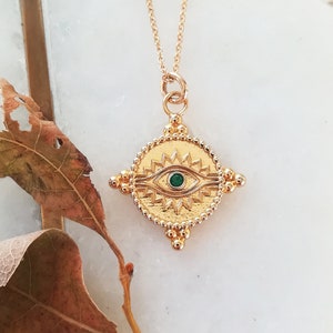 May include: A gold chain necklace with a gold pendant in the shape of a diamond with a green eye in the center. The eye is surrounded by a sunburst design.