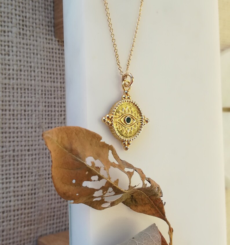 May include: A gold necklace with a square pendant featuring an evil eye design with a green stone in the center. The pendant is ornate with a beaded border and intricate details.