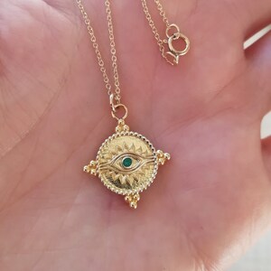 May include: A gold chain necklace with a gold pendant featuring a green eye design. The pendant is round and has a textured surface with small gold beads around the edge.