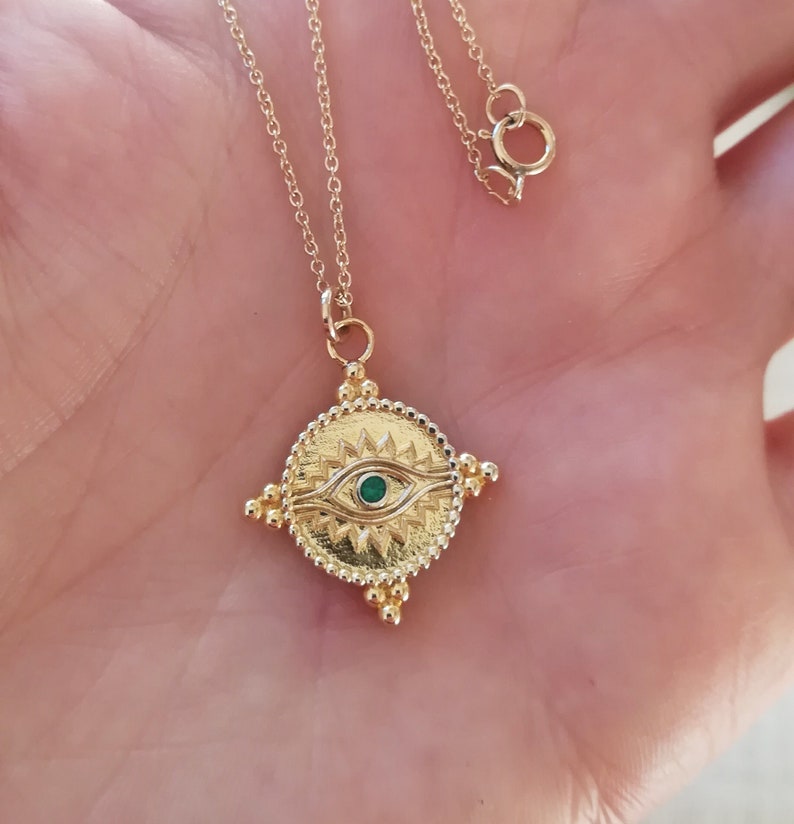 May include: A gold chain necklace with a gold pendant featuring a green eye design. The pendant is round and has a textured surface with small gold beads around the edge.