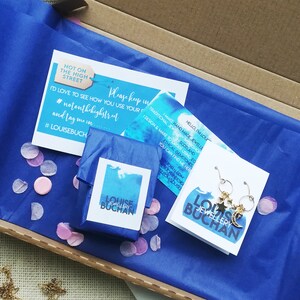 May include: A blue box with a blue tissue paper inside. The tissue paper is wrapped around a small blue box with a white label that says 'LOUISE JEWELLERY BUCHAN.' The box is surrounded by pink confetti. There is a white card with a blue watercolor design that says 'HELLO, I'M LOUISE. I MAKE HAND-MADE JEWELLERY IN A TRADITIONAL JEWELLERY WORKSHOP IN SCOTLAND. I DESIGN & MAKE TO ORDER USING ETHICAL GEMSTONES. YOU CAN FIND ME HERE: LOUISEBUCHAN.COM/LOUISEEBUCHANDESIGN.COM/HOUSE.' There is a second white card with a blue watercolor design that says 'NOT ON THE HIGH STREET. PLEASE KEEP IN TOUCH. I'D LOVE TO SEE HOW YOU USE YOUR NEW JEWELLERY. #NOTONTHEHIGHSTREET AND TAG ME IN #LOUISEBUCH.' The card is also surrounded by pink confetti. The image shows a pair of gold earrings with a star and crescent moon charm.