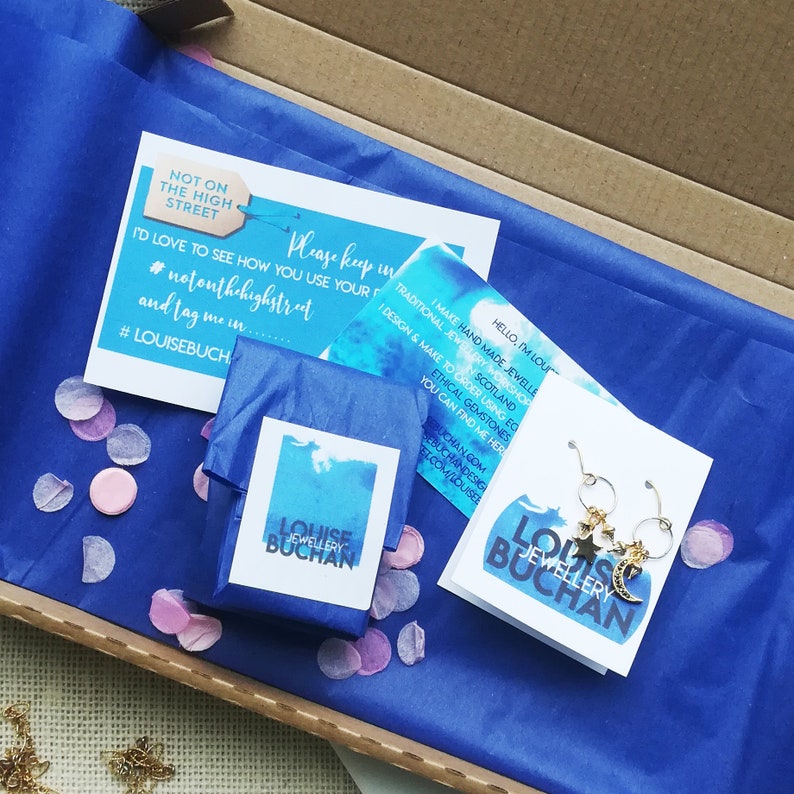 May include: A blue box with a blue tissue paper inside. The tissue paper is wrapped around a small blue box with a white label that says 'LOUISE JEWELLERY BUCHAN.' The box is surrounded by pink confetti. There is a white card with a blue watercolor design that says 'HELLO, I'M LOUISE. I MAKE HAND-MADE JEWELLERY IN A TRADITIONAL JEWELLERY WORKSHOP IN SCOTLAND. I DESIGN & MAKE TO ORDER USING ETHICAL GEMSTONES. YOU CAN FIND ME HERE: LOUISEBUCHAN.COM/LOUISEEBUCHANDESIGN.COM/HOUSE.' There is a second white card with a blue watercolor design that says 'NOT ON THE HIGH STREET. PLEASE KEEP IN TOUCH. I'D LOVE TO SEE HOW YOU USE YOUR NEW JEWELLERY. #NOTONTHEHIGHSTREET AND TAG ME IN #LOUISEBUCH.' The card is also surrounded by pink confetti. The image shows a pair of gold earrings with a star and crescent moon charm.