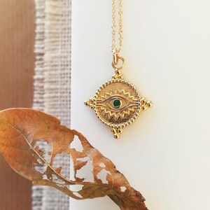May include: A gold chain necklace with a green eye charm pendant. The charm is in the shape of a diamond with a circle in the center. The circle has a gold eye with a green gemstone in the center. The eye is surrounded by gold rays.