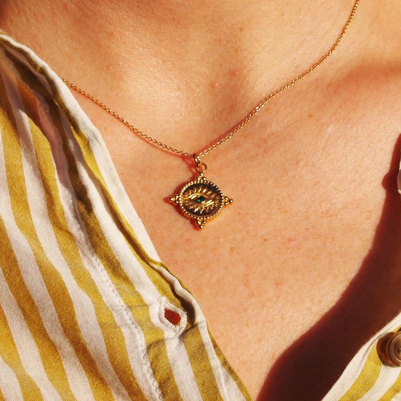 May include: A gold necklace with a pendant shaped like a four-pointed star with an eye in the center. The eye is green and the star has a textured surface.