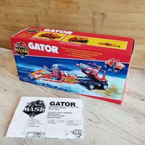 Op de afbeelding: Een rode en zwarte M.A.S.K. Gator speelgoedvoertuigdoos met een instructievel. De doos bevat illustraties van het Gator-voertuig en de tekst "Gator". Het instructievel heeft de tekst "Gator" en "Vehiculo Todo-Terreno".