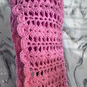 May include: A pink crocheted shawl with a scalloped edge. The openwork design reveals a darker pink fabric underneath. The shawl is made of soft yarn and is suitable for cool weather.