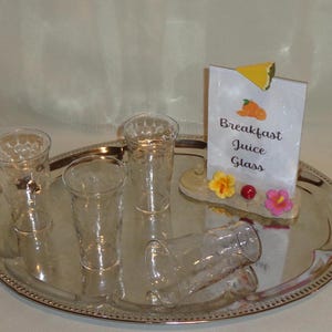 May include: Four clear glass juice glasses with a textured surface are arranged on a silver tray. A sign with the text "Breakfast Juice Glass" is on the tray with a small decorative flower arrangement.