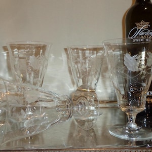 Op de afbeelding: Heldere geëtste glaswerk met bloemmotieven. De set bevat verschillende glazen, waaronder een wijnglas, een kelk en een tumbler. Een donkere wijnfles staat op de achtergrond. De glazen zijn gerangschikt op een zilveren dienblad.