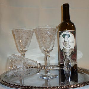 May include: A wine bottle and three etched wine glasses on a silver tray. The wine bottle has a label with a woman illustration and the word "LIBERTÉ". The glasses have a floral design.