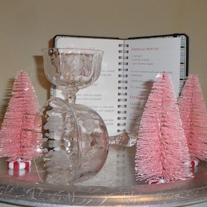 Puede incluir: Una bandeja plateada con dos copas de cóctel de cristal grabado y tres árboles de Navidad rosas de cepillo para botellas. Un libro de recetas abierto en el fondo. Los árboles tienen bases a rayas rojas y blancas.