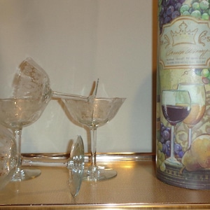 May include: Clear stemmed champagne glasses are arranged on a gold tray, with a decorative wine-themed sign in the background. The sign features images of grapes, wine glasses, and a pear.