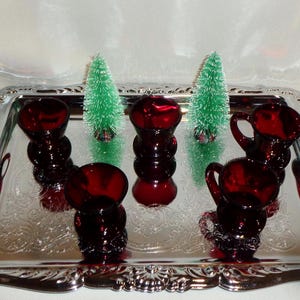 Puede incluir: Una bandeja plateada contiene varios objetos de vidrio rojo rubí, incluyendo pequeños jarrones y una jarra. También hay dos pequeños árboles de Navidad verdes en la bandeja. La bandeja tiene un borde decorativo.