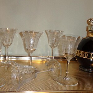 May include: A collection of etched crystal wine glasses and a black liquor bottle with a gold crown stopper. The glasses are arranged on a gold tray, with one glass lying on its side. The bottle has the words "LORD LIQUEUR" in gold.