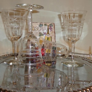 Puede incluir: Una colección de copas de vino de cristal transparente con diseños grabados, dispuestas sobre una bandeja plateada. Las copas incluyen una copa con pie, una copa de vino con un patrón de cuadrícula y una copa de vino con un diseño floral. Un letrero decorativo con la palabra "RISERVA" es visible.