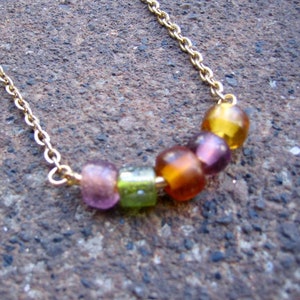 May include: A gold-toned metal chain necklace featuring five small, translucent, multi-colored beads in shades of light purple, lime green, amber, dark purple, and yellow, arranged in a horizontal row along the center of the delicate chain.