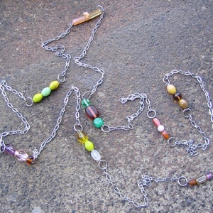 May include: A silver chain necklace with green, yellow, brown, and pink beads. The necklace is on a grey textured surface.