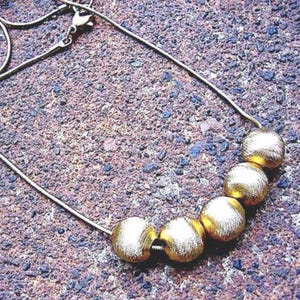 May include: A gold-colored necklace with a delicate chain and five large, textured, spherical beads. The necklace is displayed on a textured, brown and purple surface.