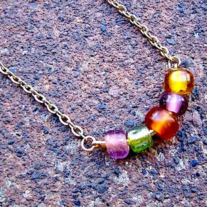 May include: A gold-toned metal chain necklace featuring a curved arrangement of five translucent, multi-colored glass beads. The beads are arranged in a sequence of purple, green, orange, dark purple, and yellow, suspended from the delicate link chain.