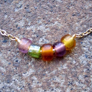 May include: A gold-toned metal chain necklace featuring five small, translucent, multi-colored glass beads in shades of purple, lime green, orange, and yellow, arranged in a row along the center of the delicate link chain.