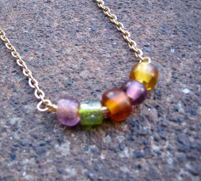 May include: A gold-toned metal chain necklace featuring five small, translucent, multi-colored beads in shades of light purple, lime green, amber, dark purple, and yellow, arranged in a horizontal row along the center of the delicate chain.