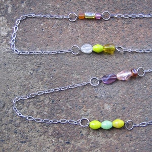 May include: Three silver chains with colourful glass beads. The top chain has orange, clear, and pink beads. The middle chain has clear, green, and yellow beads. The bottom chain has purple, pink, and green beads.