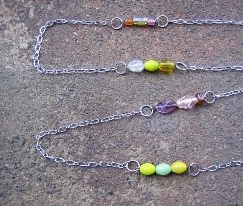 May include: Three silver chains with colourful glass beads. The top chain has orange, clear, and pink beads. The middle chain has clear, green, and yellow beads. The bottom chain has purple, pink, and green beads.