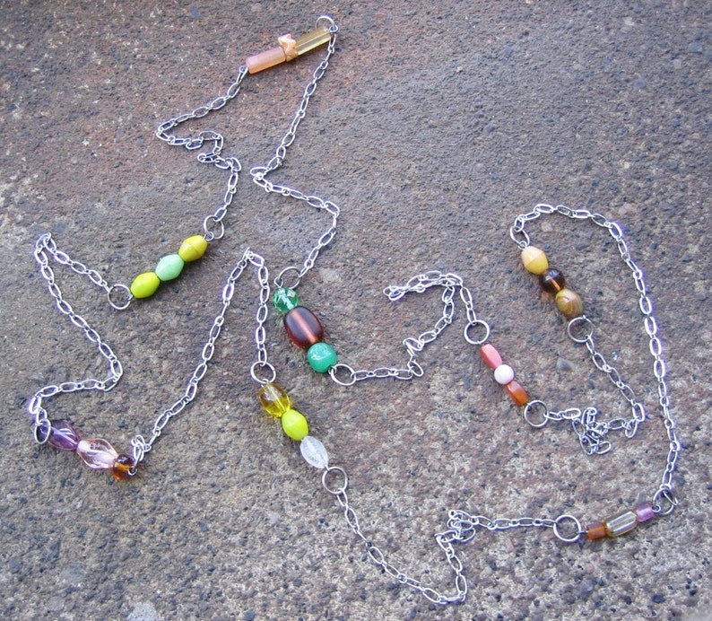 May include: A silver chain necklace with green, yellow, brown, and pink beads. The necklace is on a grey textured surface.
