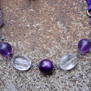 K&ouml;nnte beinhalten: Eine Halskette aus violetten und klaren Glasperlen mit einer silbernen Kette und violetten Schleifenb&auml;ndern.