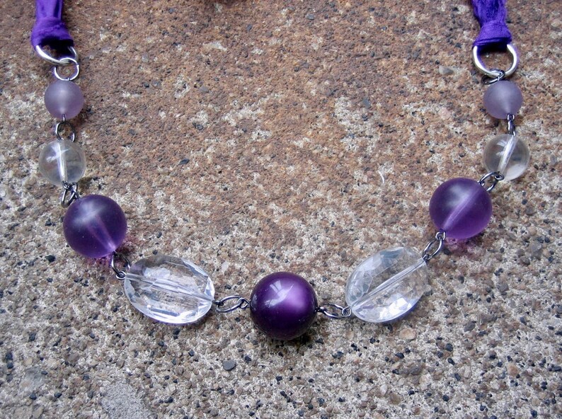 K&ouml;nnte beinhalten: Eine Halskette aus violetten und klaren Glasperlen mit einer silbernen Kette und violetten Schleifenb&auml;ndern.