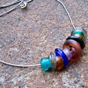May include: A silver chain necklace with a colorful glass bead pendant. The pendant is made of several stacked glass beads in shades of blue, green, orange, and purple.