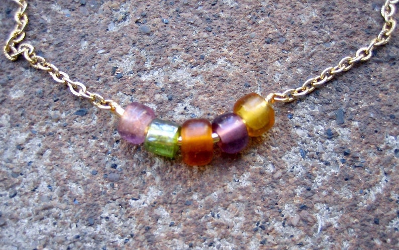 May include: A gold-toned metal chain necklace featuring five small, translucent, multi-colored glass beads in shades of purple, lime green, orange, and yellow, arranged in a row along the center of the delicate link chain.