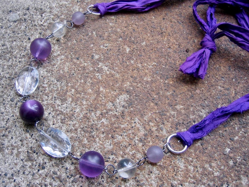 K&ouml;nnte beinhalten: Eine violette und klare Glasperlenkette mit einer violetten Schleife. Die Kette besteht aus abwechselnd klaren und violetten runden Perlen.