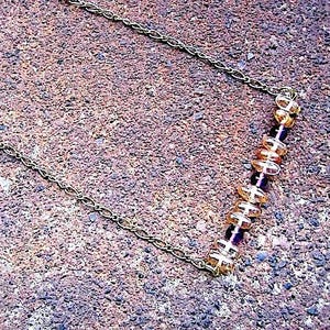 May include: A delicate necklace featuring a thin gold-toned metal chain. The center section is adorned with a linear arrangement of alternating translucent amber-colored beads and smaller, dark purple spherical beads, creating a simple pattern.