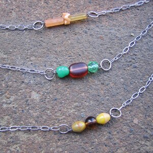 May include: Three silver chains with different coloured beads. The top chain has a light brown and orange bead. The middle chain has a green, brown, and green bead. The bottom chain has a yellow, brown, and green bead.