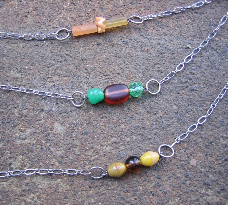 May include: Three silver chains with different coloured beads. The top chain has a light brown and orange bead. The middle chain has a green, brown, and green bead. The bottom chain has a yellow, brown, and green bead.