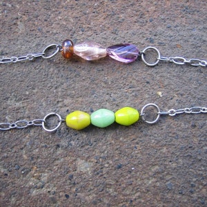 May include: Two silver chains with different coloured glass beads. The top chain has brown, pink and purple beads. The bottom chain has yellow and green beads.