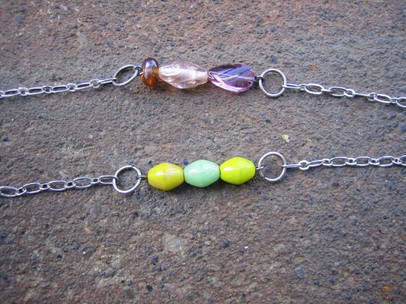 May include: Two silver chains with different coloured glass beads. The top chain has brown, pink and purple beads. The bottom chain has yellow and green beads.
