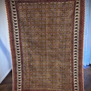 May include: A rectangular, patterned textile with a brown background and repeating floral and geometric designs. The border features a paisley pattern in black and white, with a red and white trim. The textile has a fringe along the top and bottom edges.