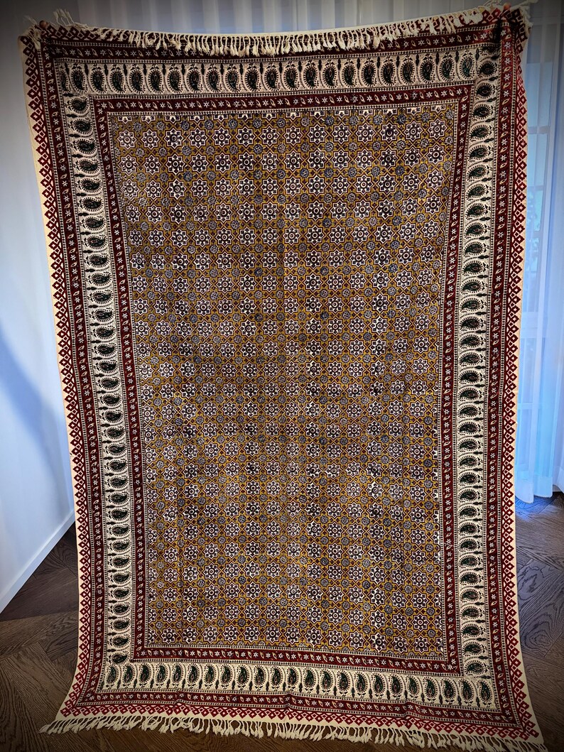 May include: A rectangular, patterned textile with a brown background and repeating floral and geometric designs. The border features a paisley pattern in black and white, with a red and white trim. The textile has a fringe along the top and bottom edges.