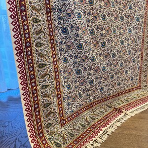 May include: A rectangular, patterned rug with a cream-colored background. The rug features a repeating floral design in shades of blue, pink, and green. The border is a deep red with geometric and paisley patterns, and the rug has a fringed edge.